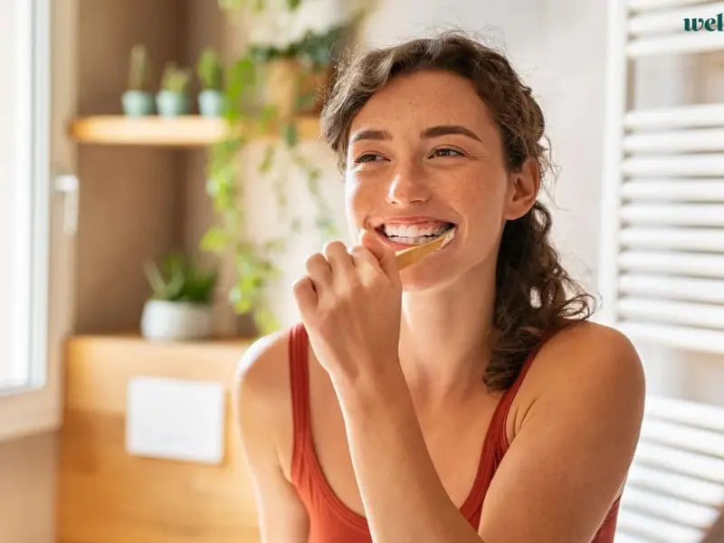 Enamel-safe toothpaste used by a smiling woman brushing teeth with a bamboo toothbrush in a bright bathroom.