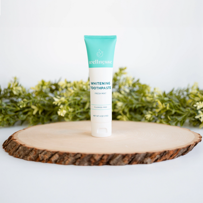 All-natural whitening toothpaste tube labeled Wellnesse, displayed on a wood slice with greenery in the background.