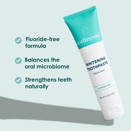 Nontoxic whitening toothpaste in a Wellnesse tube, displayed on a wood slab with green foliage in the background.