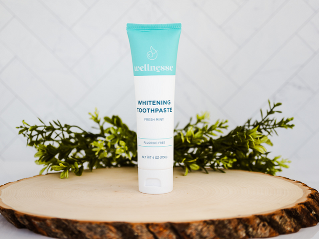 Fluoride free whitening toothpaste displayed on a wooden slab with greenery, featuring a Wellnesse fresh mint tube.