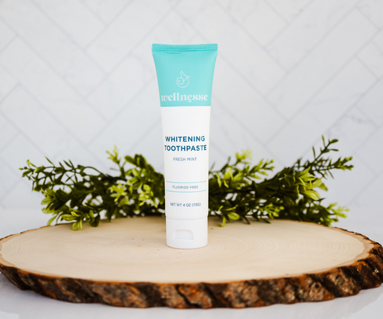 Fluoride free whitening toothpaste displayed on a wooden slab with greenery, featuring a Wellnesse fresh mint tube.