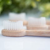 Bamboo toothbrush with soft white bristles shown in close-up on a glossy white surface with blurred greenery in the background