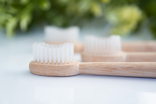 Bamboo toothbrush with soft white bristles shown in close-up on a glossy white surface with blurred greenery in the background