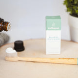 Concentrated mouthwash being dropped onto toothpaste-covered bamboo toothbrush next to Wellnesse packaging and natural decor.