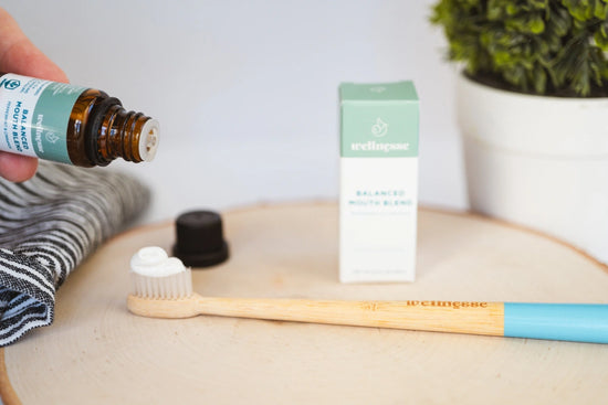 Concentrated mouthwash being dropped onto toothpaste-covered bamboo toothbrush next to Wellnesse packaging and natural decor.