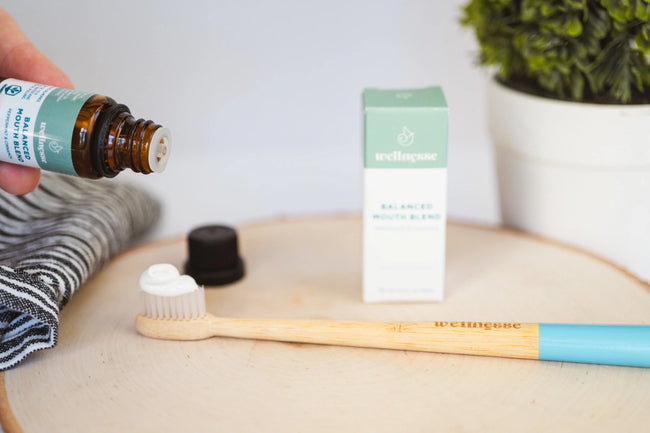 Concentrated mouthwash being dropped onto toothpaste-covered bamboo toothbrush next to Wellnesse packaging and natural decor.