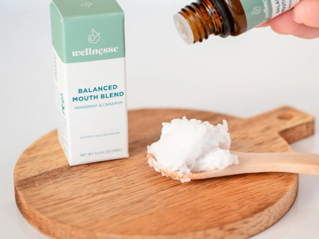 Concentrated mouthwash being dropped onto a spoonful of coconut oil next to Wellnesse Balanced Mouth Blend box.