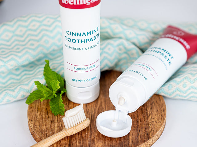 Cinnamon toothpaste without fluoride in a Wellnesse Cinnamint tube, shown with bamboo toothbrush and fresh mint on a wooden tray.
