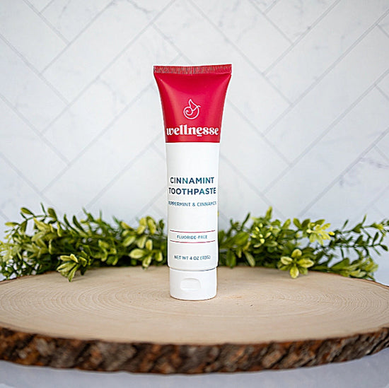 Cinnamon toothpaste by Wellnesse in a red and white tube, displayed upright on wood slice with greenery background.