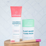 Strawberry chemical free toothpaste for children by Wellnesse, paired with plant-based floss picks and a pink bamboo toothbrush.