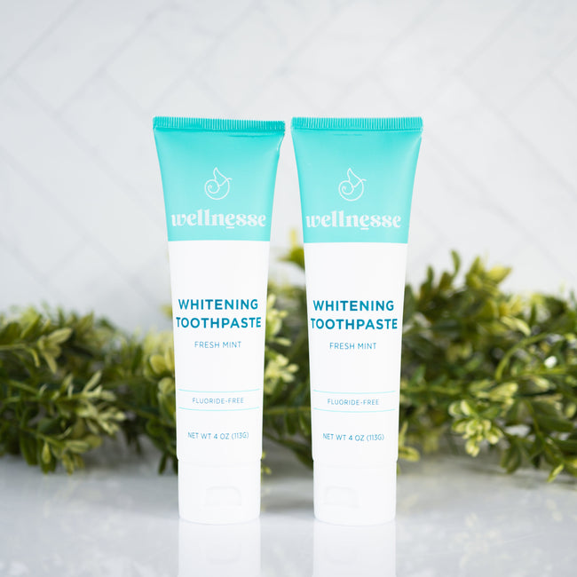 All natural whitening toothpaste tubes from Wellnesse, featuring fresh mint flavor and fluoride-free formula with green foliage background.
