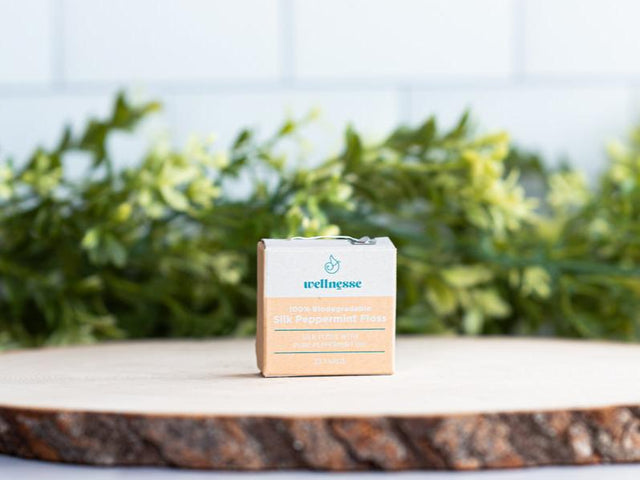 Silk dental floss in biodegradable packaging displayed on a wood slice with green foliage in the background