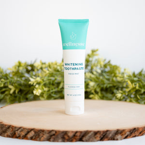 Non toxic whitening toothpaste in a Wellnesse fresh mint tube, displayed on a wooden slab with green leafy background.
