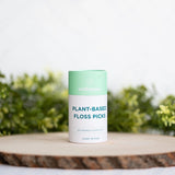 Floss picks in a mint green and white Wellnesse container labeled plant-based, displayed on a wooden surface with greenery.