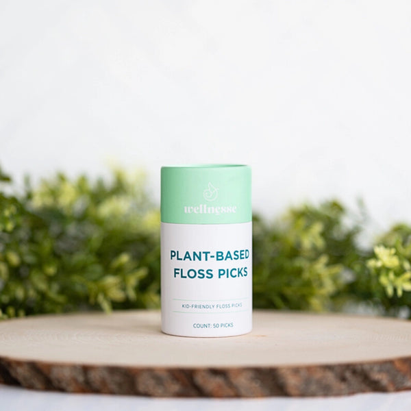 Floss picks in a mint green and white Wellnesse container labeled plant-based, displayed on a wooden surface with greenery.