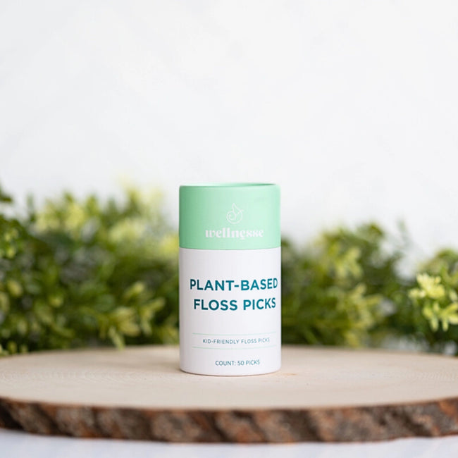 Floss picks in a mint green and white Wellnesse container labeled plant-based, displayed on a wooden surface with greenery.