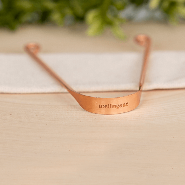 Copper tongue scraper with "Wellnesse" engraved, resting on a light cloth with greenery in the background.