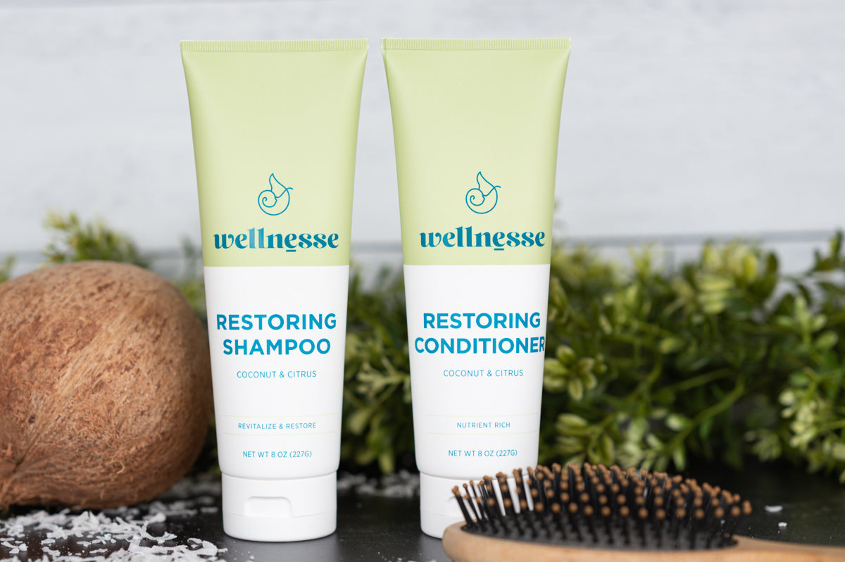 Wellnesse restoring shampoo and conditioner tubes beside a coconut and wooden hairbrush.