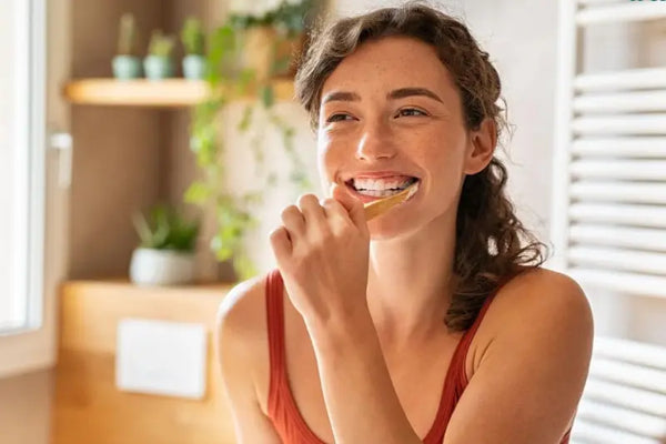 Enamel-safe toothpaste used by a smiling woman brushing teeth with a bamboo toothbrush in a bright bathroom.