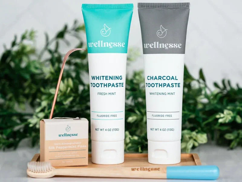 Non-abrasive charcoal toothpaste and whitening toothpaste tubes displayed with floss and bamboo toothbrush on a wooden tray.