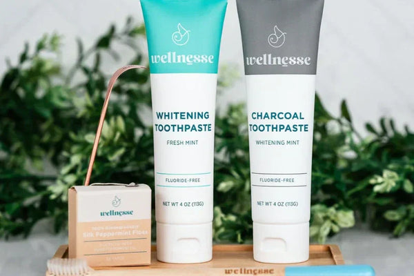 Non-abrasive charcoal toothpaste and whitening toothpaste tubes displayed with floss and bamboo toothbrush on a wooden tray.