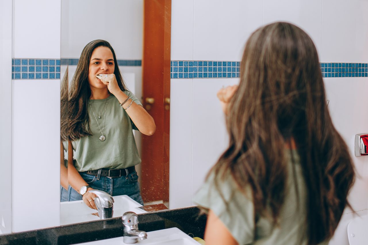 Woman brushing teeth in bathroom mirror.