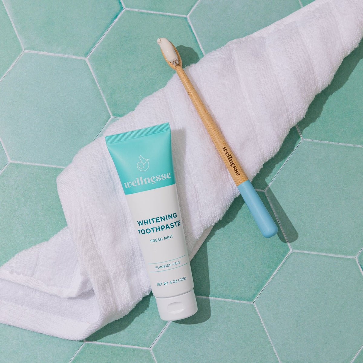 Wellnesse Whitening Toothpaste with a bamboo toothbrush on a white towel over teal tile.