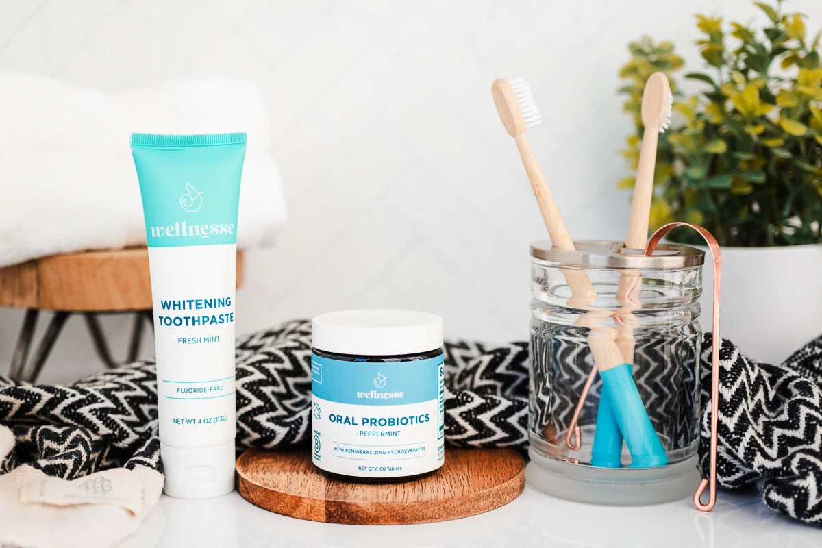 Natural toothpaste with hydroxyapatite from Wellnesse shown alongside oral probiotics, bamboo toothbrushes, and a copper tongue scraper on a bathroom counter.