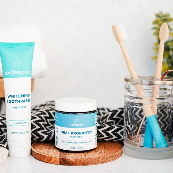 Natural toothpaste with hydroxyapatite from Wellnesse shown alongside oral probiotics, bamboo toothbrushes, and a copper tongue scraper on a bathroom counter.