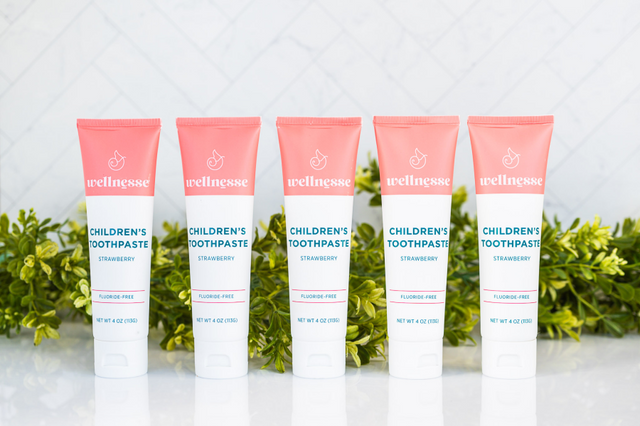Strawberry healthy natural toothpaste in five fluoride-free tubes lined up with green foliage on a reflective white surface