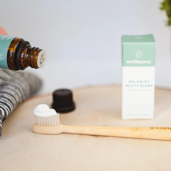 Concentrated mouthwash being dropped onto toothpaste-covered bamboo toothbrush next to Wellnesse packaging and natural decor.