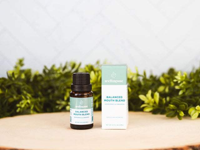 Concentrated mouthwash bottle and box labeled Balanced Mouth Blend with peppermint and cinnamon, displayed on wood with greenery.