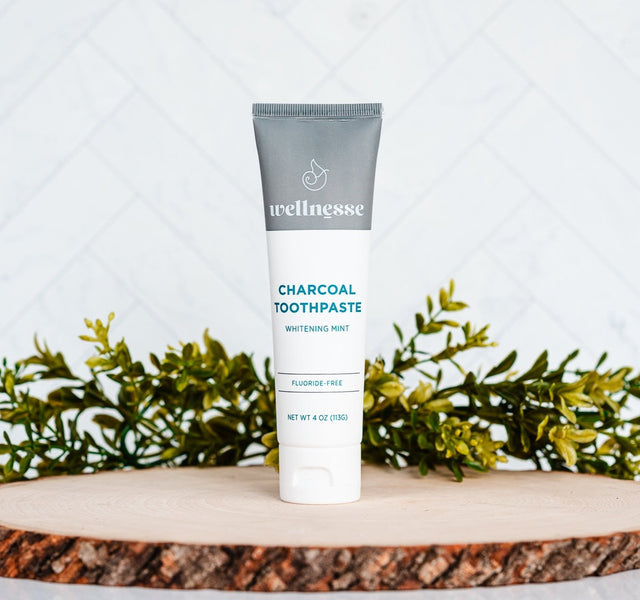Wellnesse charcoal toothpaste tube standing on a rustic wood display with fresh greenery in the background