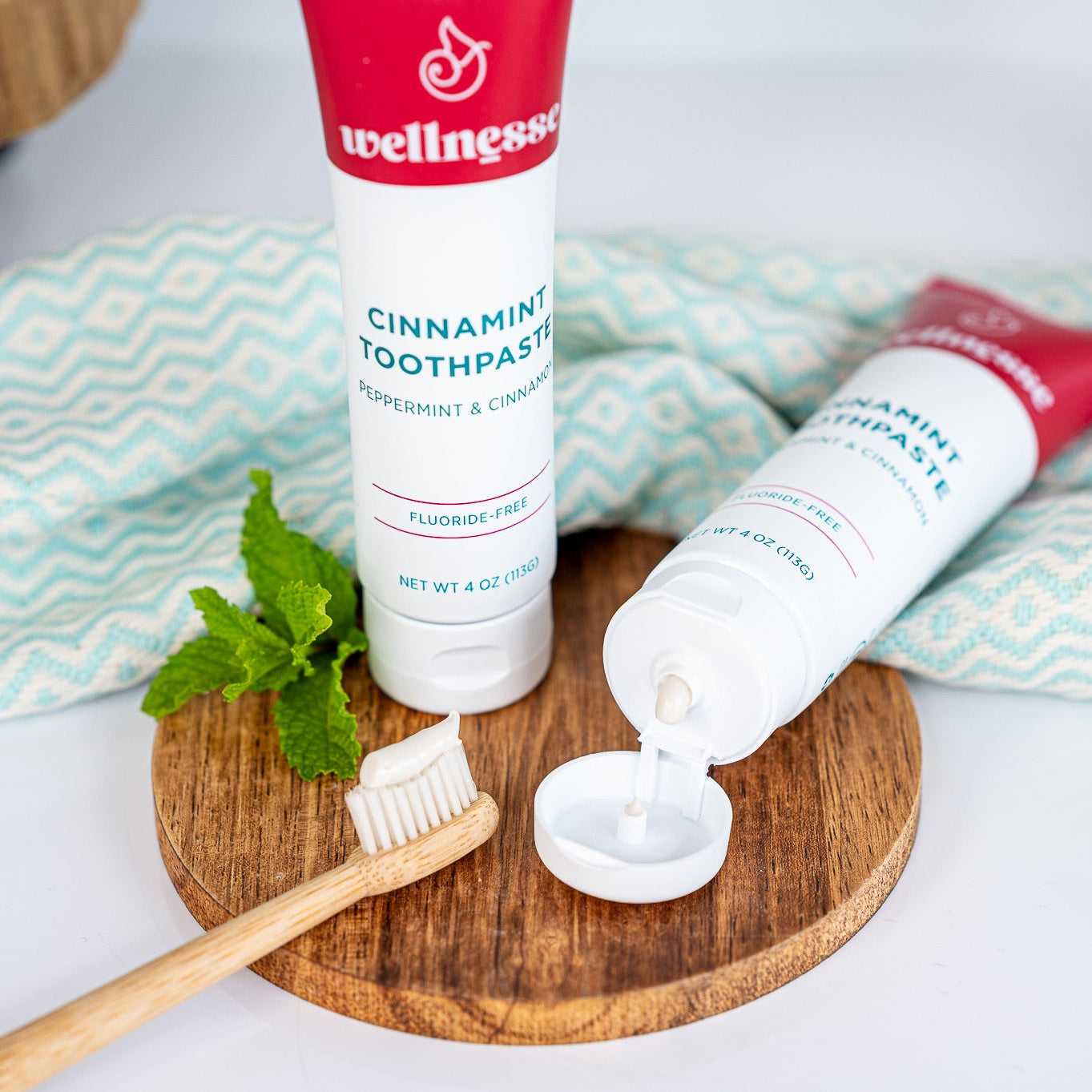 Cinnamon toothpaste without fluoride in a Wellnesse Cinnamint tube, shown with bamboo toothbrush and fresh mint on a wooden tray.