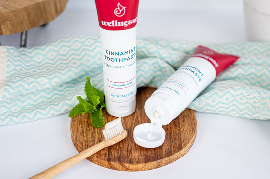 Cinnamon toothpaste without fluoride in a Wellnesse Cinnamint tube, shown with bamboo toothbrush and fresh mint on a wooden tray.