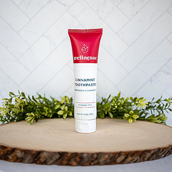 Wellnesse cinnamon toothpaste in a red and white tube on a wood slice with greenery behind
