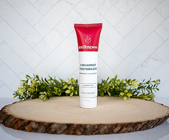 Cinnamon toothpaste by Wellnesse in a red and white tube, displayed upright on wood slice with greenery background.