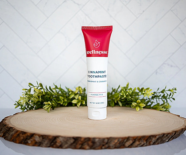 Cinnamon toothpaste by Wellnesse in a red and white tube, displayed upright on wood slice with greenery background.