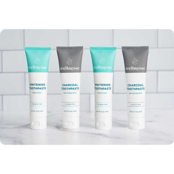 Lineup of four Wellnesse natural toothpaste tubes alternating fresh mint whitening and charcoal whitening mint flavors on a marble counter.
