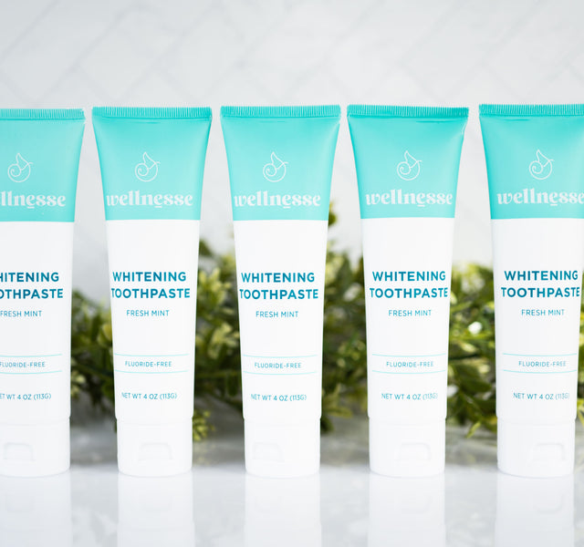 Mint whitening toothpaste five-pack from Wellnesse with fluoride-free fresh mint formula, displayed in front of lush green leaves.