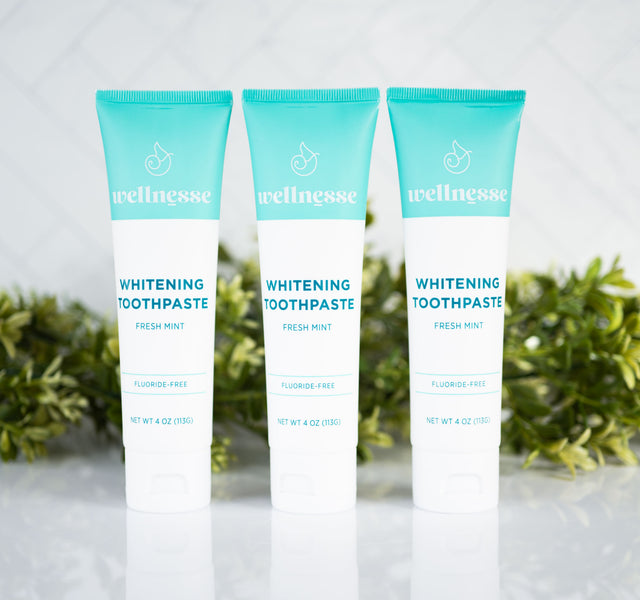Organic whitening toothpaste trio in Wellnesse fluoride-free fresh mint formula, displayed with green foliage on a tiled surface.