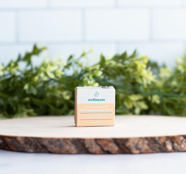 Wellnesse silk dental floss in biodegradable packaging on a wood slice with green foliage behind