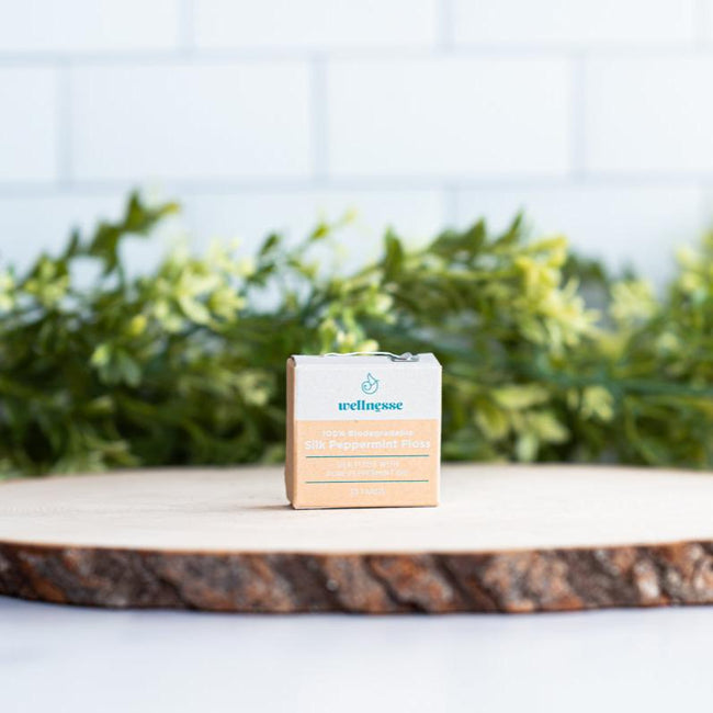 Silk dental floss in biodegradable packaging displayed on a wood slice with green foliage in the background