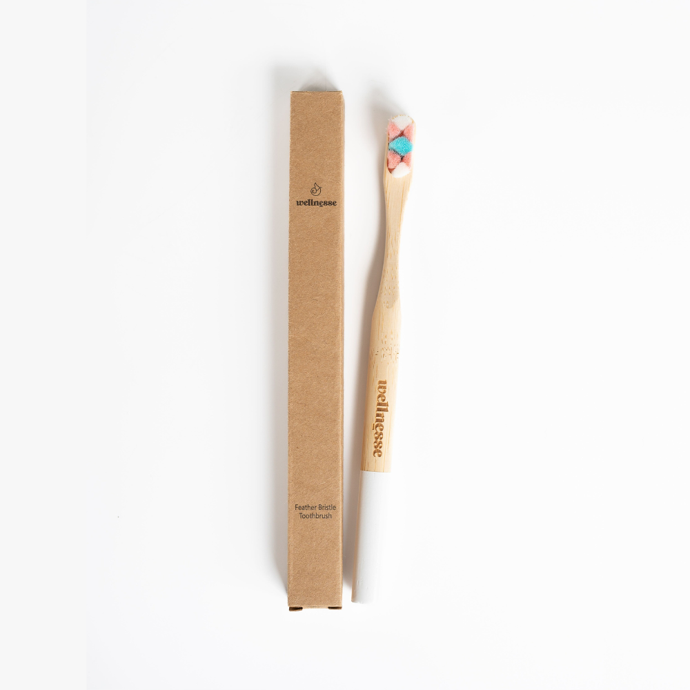 Bamboo toothbrush with soft feather bristles in pink and blue, shown beside its recyclable Wellnesse packaging.