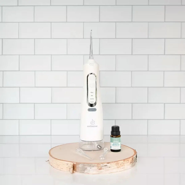 Wellnesse water flosser paired with Balanced Mouth Blend oil on a natural wood slice against white tile