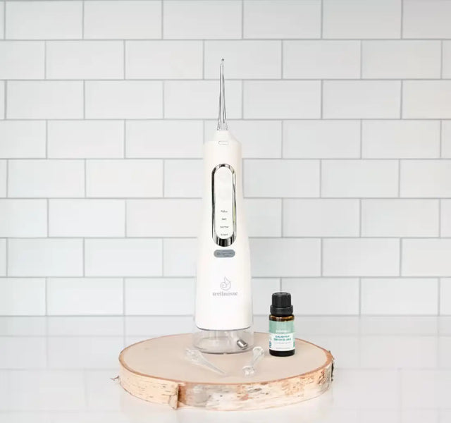 Wellnesse water flosser paired with Balanced Mouth Blend oil on a natural wood slice against white tile