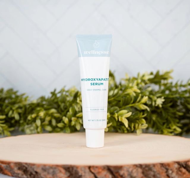 Wellnesse hydroxyapatite serum fluoride-free tube on a natural wood slice with green foliage backdrop
