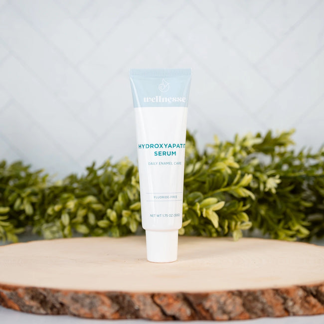 Wellnesse Hydroxyapatite Serum fluoride-free tube standing on a natural wood slice with green foliage backdrop.