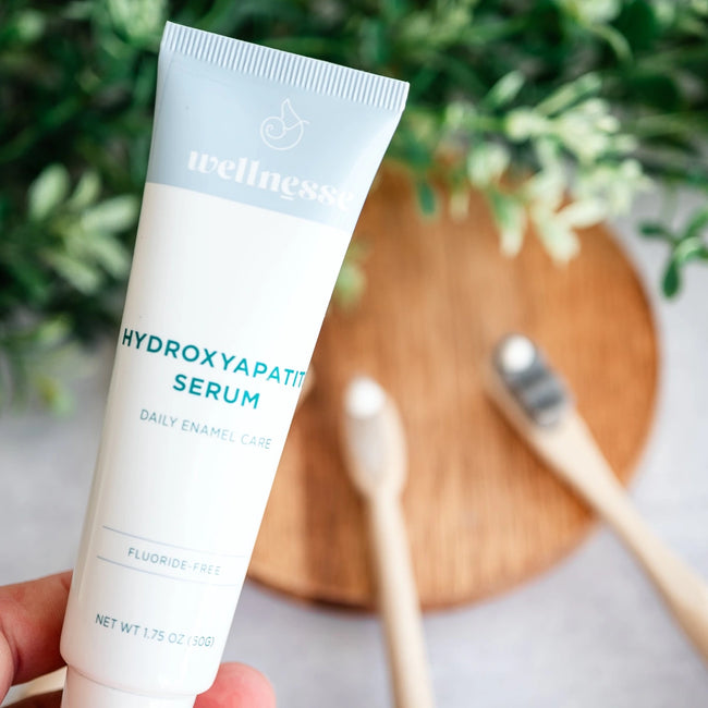 Close-up of Wellnesse Hydroxyapatite Serum fluoride-free tube held in hand with a bamboo toothbrush and wooden tray behind.