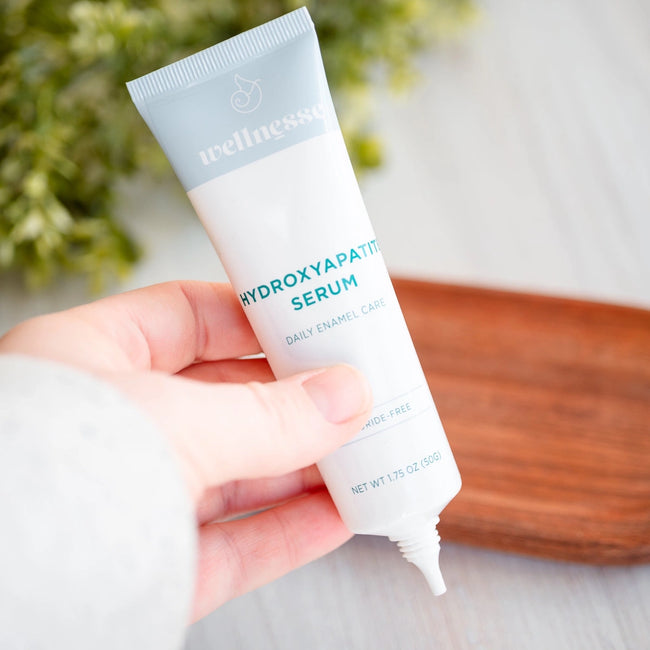 Hand holding a tube of Wellnesse Hydroxyapatite Serum for daily enamel care with greenery in the background.